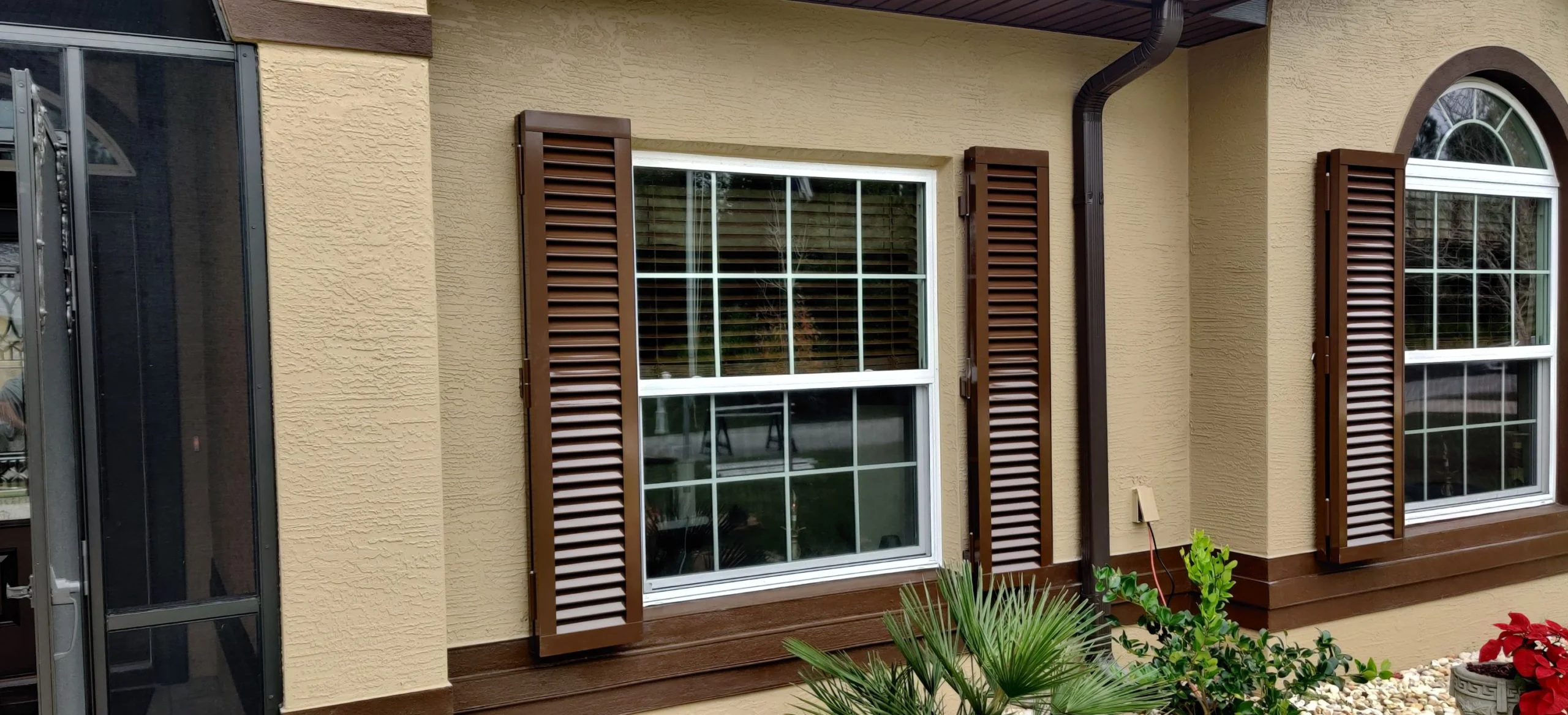 A double-hung window with brown shutters is mounted on a beige stucco wall beside another window with an arched top