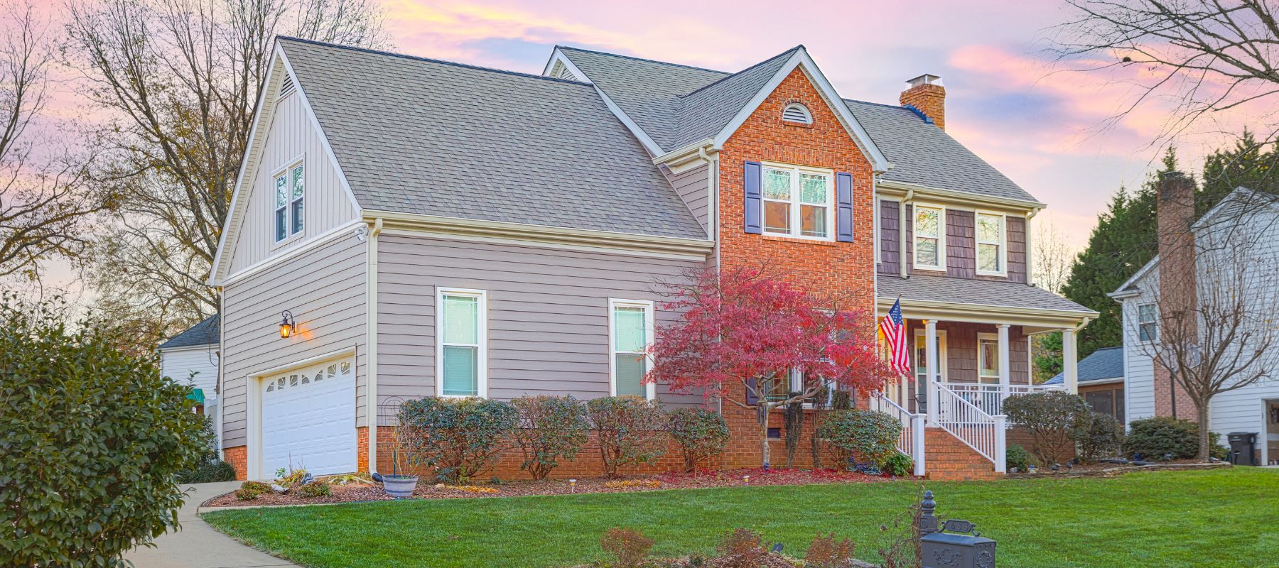 A large, two-story house with a mix of gray siding and brick facade stands on a green lawn under a colorful sky at dusk, with an American flag by the porch