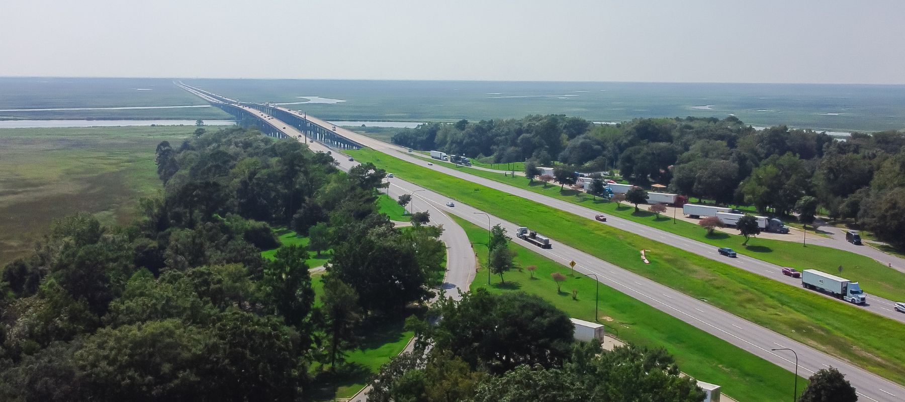 An aerial view shows a multi-lane highway and a long bridge stretching across wetlands and green trees under a clear sky