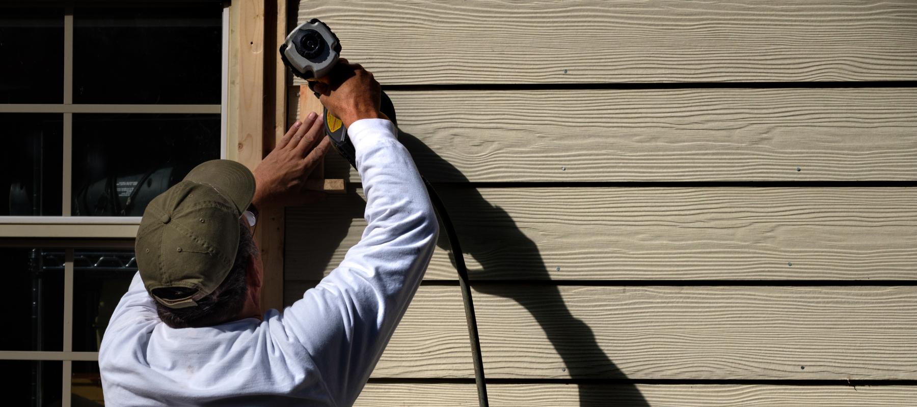 A contractor using a power tool to install beige horizontal siding on a house exterior